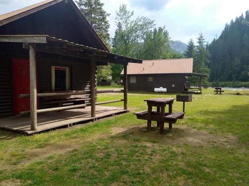 Two rustic cabins with wooden porches and picnic tables in a grassy area surrounded by trees and mountains.