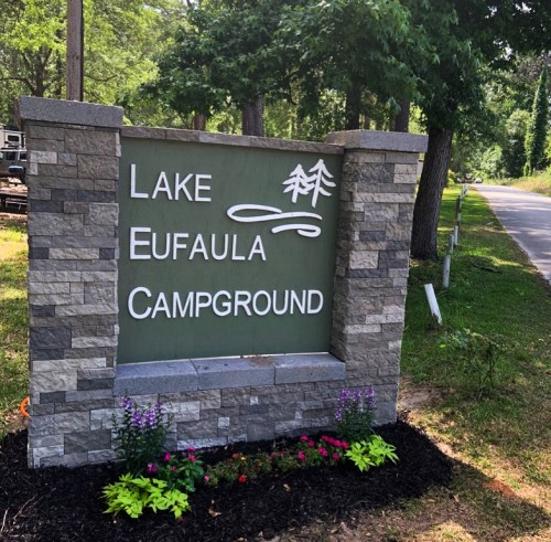 Sign for Lake Eufaula Campground, featuring trees and flowers, set against a green background.