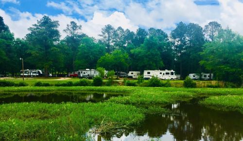 A serene campsite with RVs parked near a pond, surrounded by lush greenery and trees under a partly cloudy sky.