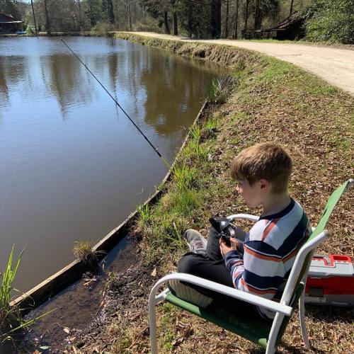 A child sits on a chair by a pond, fishing with a rod, surrounded by trees and a dirt path.