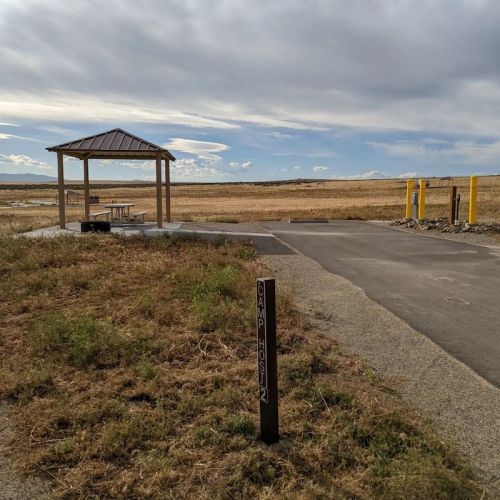 A picnic shelter near a paved path in an open field under a cloudy sky, with a sign for "Camp Host 12."