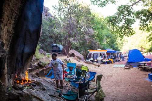 A child stands near a small fire in a campsite surrounded by tents and camping chairs in a wooded area.