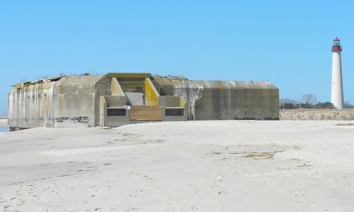 A weathered concrete bunker on a sandy beach, with a lighthouse in the background under a clear blue sky.