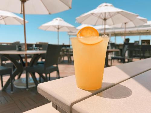 A refreshing drink with a lemon slice sits on a table, with umbrellas and outdoor seating in the background.