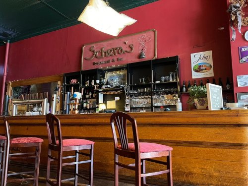Interior of a restaurant bar with wooden counter, empty chairs, and a red wall featuring a sign for "Scher's Restaurant & Bar."