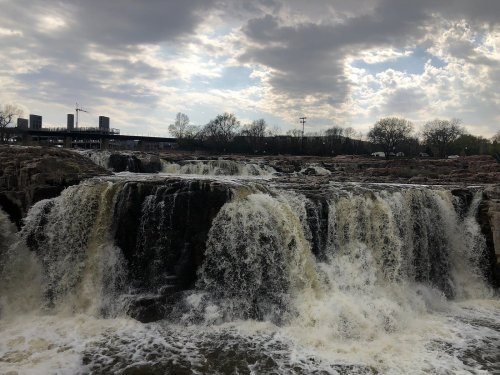 A waterfall cascades over rocky cliffs under a cloudy sky, with trees and distant buildings in the background.