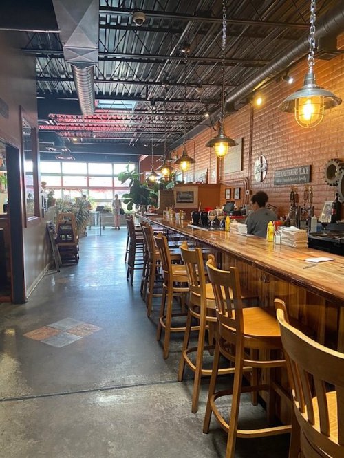 A cozy restaurant interior featuring a long wooden bar, high stools, and warm lighting along a brick wall.
