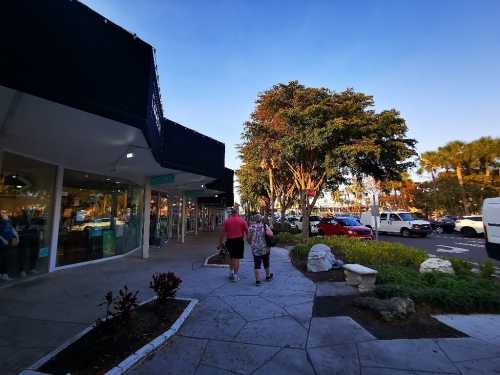 A couple walks along a sidewalk lined with shops and trees under a clear blue sky.