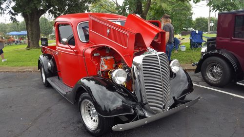 A classic red and black vintage truck with an open hood, showcasing its engine, parked at a car show.