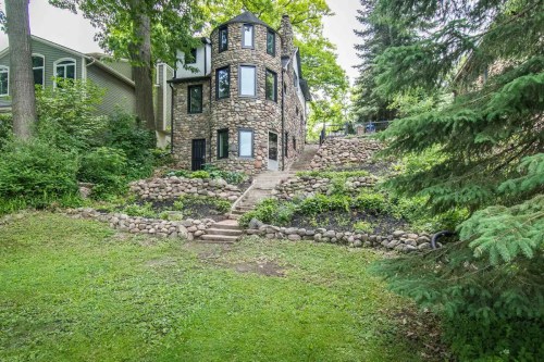 A unique stone house surrounded by greenery and trees, with a landscaped yard and stone steps leading up.