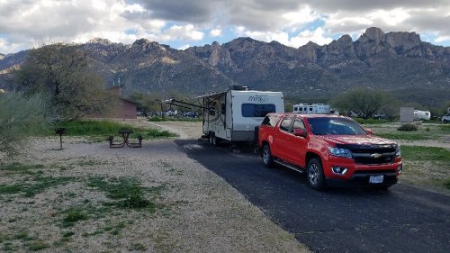 A red truck parked next to a camper trailer in a scenic area with mountains in the background and a cloudy sky.