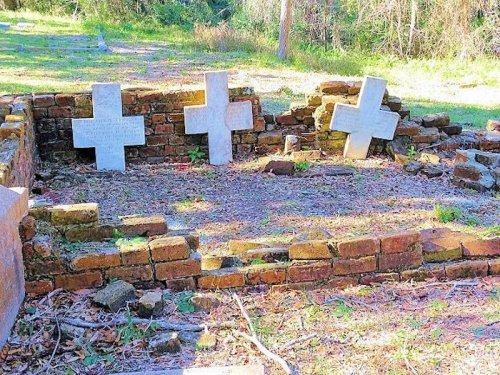 Three weathered crosses stand among crumbling brick ruins in a grassy area, surrounded by trees.