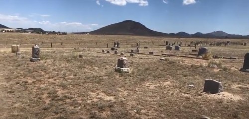 A vast, dry landscape with scattered gravestones and a distant mountain under a clear blue sky.