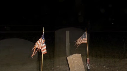 Two tattered American flags stand beside a gravestone in a dark, quiet cemetery at night.