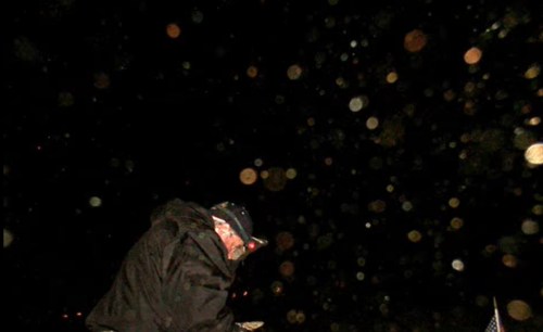 A person in a dark jacket bends down, surrounded by blurred orbs of light against a black background.