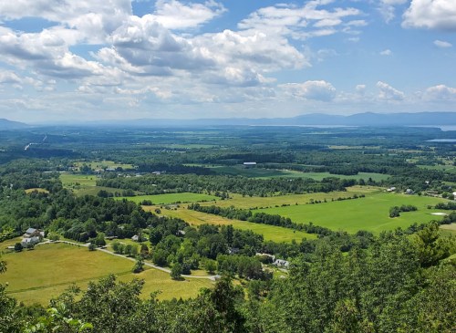 A panoramic view of lush green fields and distant mountains under a partly cloudy sky.