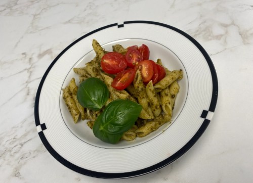 A plate of penne pasta with pesto, topped with cherry tomatoes and fresh basil leaves on a marble surface.