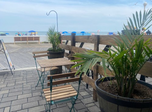 A beachside view with wooden seating, potted plants, and umbrellas in the background under a cloudy sky.