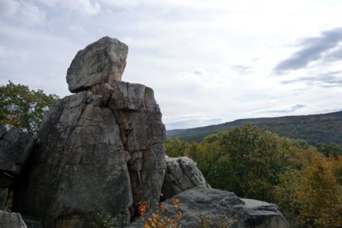 A large, balanced rock sits atop a rugged formation, overlooking a scenic landscape of trees and distant hills under a cloudy sky.