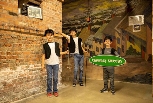 Three children dressed as chimney sweeps pose in a brick-walled room, one holding a broom and another a sign.