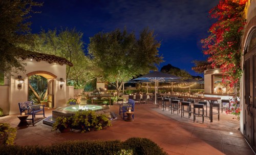 A beautifully lit outdoor patio with a fountain, seating, and vibrant flowers at night.