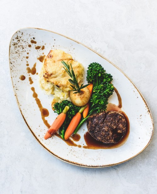 A plated meal featuring a meat patty, mashed potatoes, roasted carrots, and broccoli, drizzled with sauce.