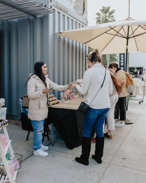 A vendor hands a product to a customer at an outdoor market, with others browsing nearby under an umbrella.