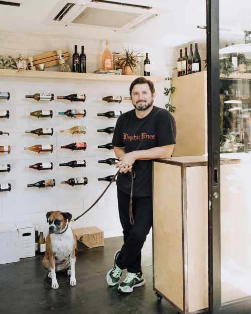 A man stands with a dog in a wine shop, surrounded by bottles on the wall and a wooden counter.