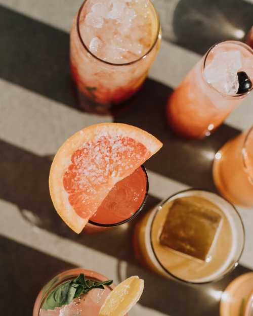 A variety of colorful cocktails with ice, garnished with fruit slices, arranged on a table with shadows.