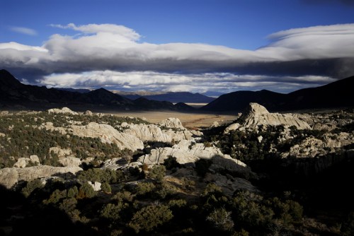 A dramatic landscape featuring rocky formations, rolling hills, and a cloudy sky, with distant mountains in the background.
