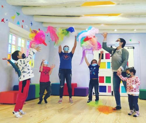 Children and adults play with colorful scarves in a bright, cheerful room, all wearing masks and smiling.