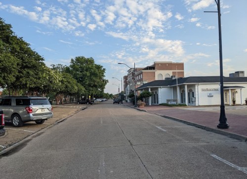 A quiet street lined with trees, parked cars, and buildings under a blue sky with scattered clouds.
