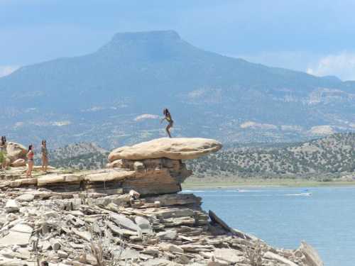 A person stands on a large rock by a lake, with mountains in the background and others nearby enjoying the scenery.