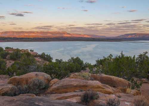 A serene landscape featuring a calm lake surrounded by rocky terrain and distant mountains under a colorful sky.