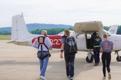 Four people in jump suits walk towards a small airplane on a runway, preparing for a skydiving jump.