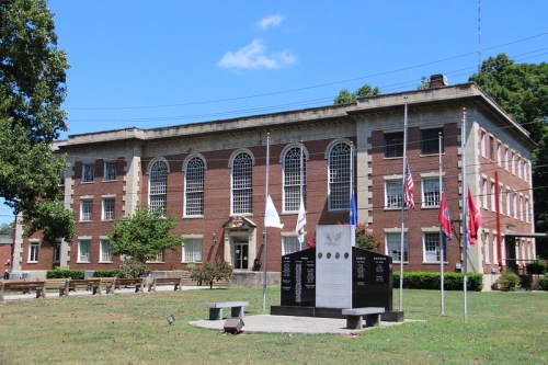 Historic brick building with large windows, surrounded by flags and a memorial in a grassy area. Clear blue sky above.