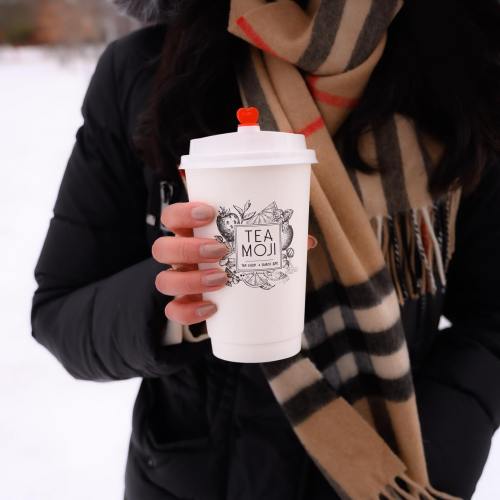 A person holding a cup with a "Tea Moji" logo, wearing a scarf and a black jacket, outdoors in a chilly setting.