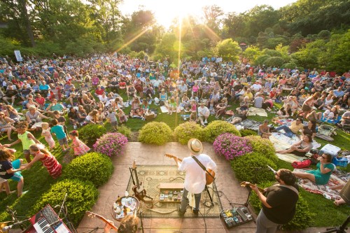 A large crowd enjoys an outdoor concert in a park, with musicians performing on stage as the sun sets.