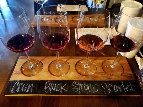 A wooden tasting board with four wine glasses filled with different red wines, labeled "Chon," "Black," "Straw," and "Scarlet."