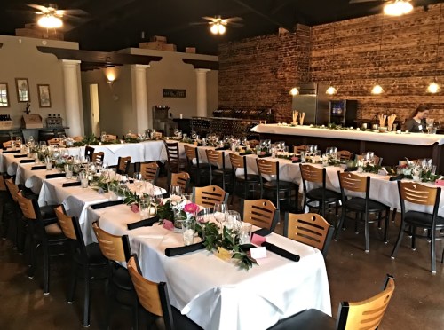 A spacious restaurant set for a formal event, with tables adorned in white linens and floral centerpieces.