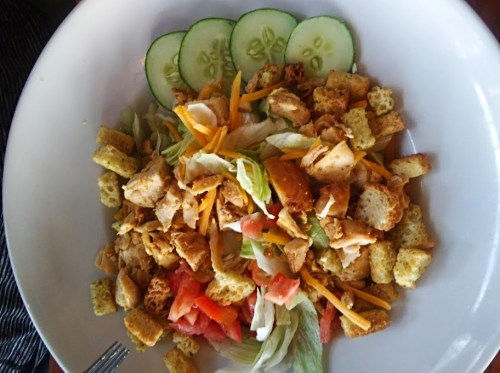 A fresh salad with grilled chicken, lettuce, tomatoes, croutons, shredded cheese, and cucumber slices.