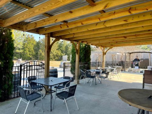 Outdoor seating area with tables and chairs under a wooden pergola, surrounded by greenery and a gravel path.
