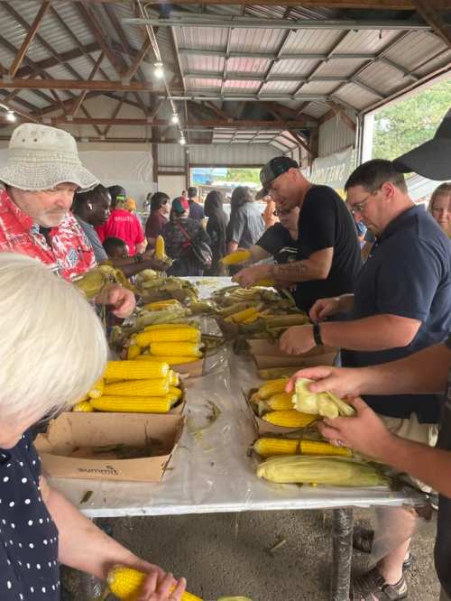 A group of people shucking corn at a long table in a barn-like setting, with others in the background.