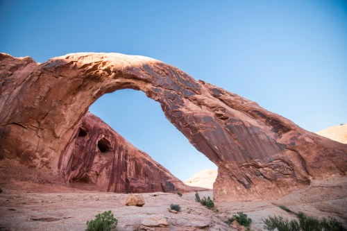A natural rock arch stands against a clear blue sky, surrounded by rugged red rock formations.