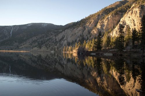 A serene lake reflecting mountains and trees at sunset, with calm water and a clear sky.