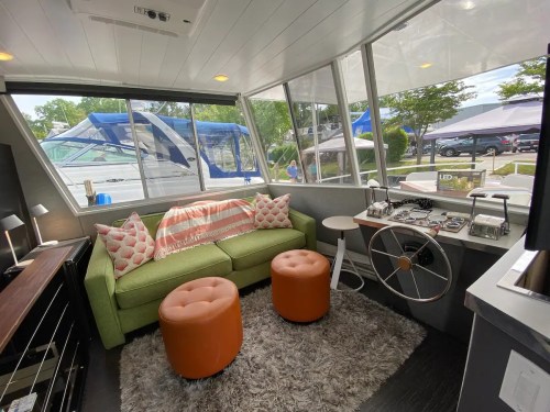 Cozy interior of a boat with a green sofa, orange stools, and large windows overlooking a marina.