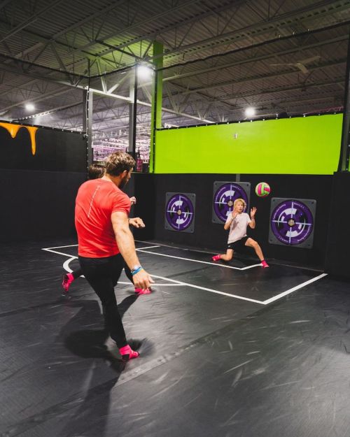A man kicks a ball towards a child in a trampoline park, both engaged in a playful game. Bright colors and fun atmosphere.