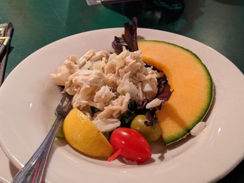 A plate of salad with shredded chicken, mixed greens, lemon, grapes, cherry tomato, and a slice of cantaloupe.