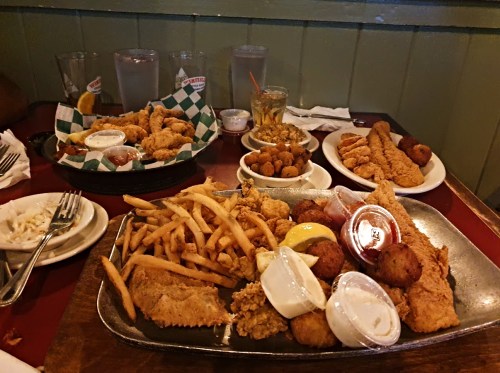 A table spread with fried fish, chicken tenders, fries, hushpuppies, and dipping sauces, alongside drinks.