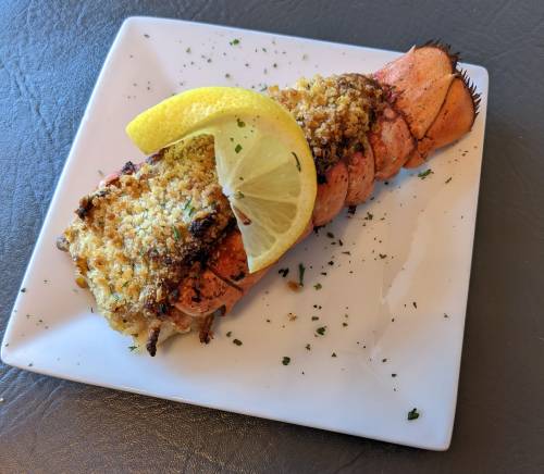 A baked lobster tail topped with breadcrumbs and a lemon wedge, garnished with parsley on a white plate.